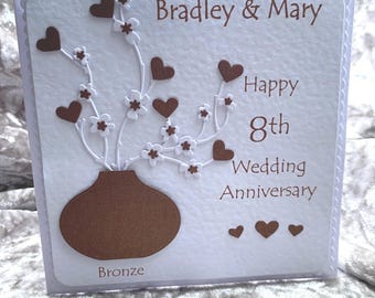 May include: A handmade 8th wedding anniversary card. The card features a brown vase with white flowers and brown hearts. The text reads "Bradley & Mary", "Happy 8th Wedding Anniversary", and "Bronze".