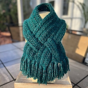 May include: A teal green crocheted scarf with fringe on the ends. The scarf is displayed on a wooden stand.