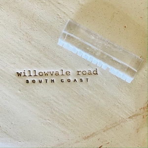 May include: A transparent plastic ruler with a notched edge lies on a textured, beige surface. Below, the words "willowvale road SOUTH COAST" are spelled out in small, brown letters. The image has a neutral, earthy tone.