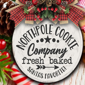 May include: A white round sign with the text "North Pole Cookie Company" in black lettering. The sign also has the text "fresh baked" and "Santa's Favorite!" in black lettering. The sign is decorated with a red and black plaid bow and a sprig of holly.