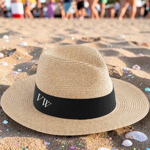 Puede incluir: Un sombrero fedora beige con ala ancha y banda negra. La banda muestra las letras blancas "VW". El sombrero está sobre una playa de arena, evocando el verano o las vacaciones.