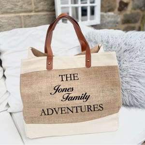 May include: A beige and white tote bag with brown leather handles. The bag features the text "THE Jones Family ADVENTURES" in black script. The bag is made of a natural fiber material.