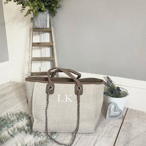 May include: A large beige canvas tote bag with brown leather trim and a silver chain strap. The bag has the initials "LK" embroidered on the front.