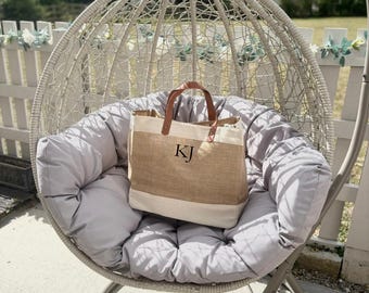 May include: A beige and white tote bag with brown leather handles and the initials "KJ" printed on the front. The bag is sitting on a gray cushion inside a white wicker hanging chair, with a white fence in the background.
