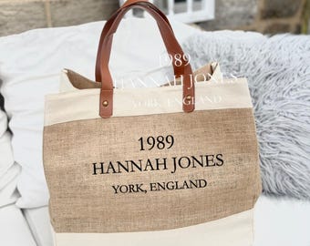 May include: A beige and brown tote bag with brown leather handles. The bag features the text "1989 HANNAH JONES YORK, ENGLAND" in black. The bag is made of a canvas-like material.
