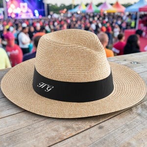 Puede incluir: Un sombrero fedora marrón claro con ala ancha y banda negra. La banda presenta las iniciales blancas "HG". El sombrero está hecho de material tejido y descansa sobre una superficie de madera.