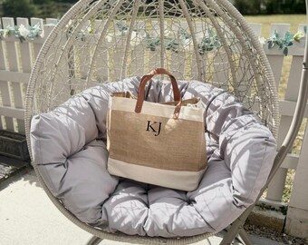 May include: A beige and white tote bag with brown leather handles and the initials "KJ" printed on the front. The bag is sitting on a gray cushion inside a white wicker hanging chair, with a white fence in the background.