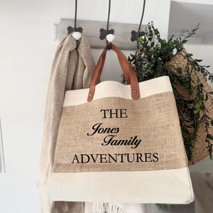 May include: A beige and cream tote bag with brown leather handles hangs from a hook. The bag has the text "THE Jones Family ADVENTURES" in black. A beige scarf hangs on a hook to the left, and greenery is visible to the right.