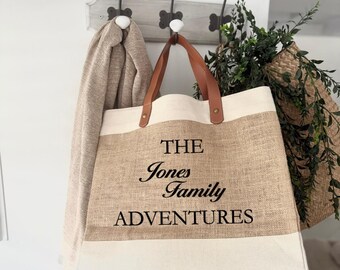 May include: A beige and white tote bag with brown leather handles hangs from a hook. The bag features the text "THE Jones Family ADVENTURES" in black. A beige scarf hangs on a hook to the left, and greenery is visible to the right.