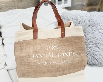 May include: A beige and white tote bag with brown leather handles. The bag features the text "1989 HANNAH JONES YORK ENGLAND" in white. The bag is made of a woven material and has a rectangular shape.