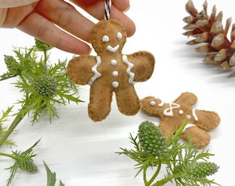 May include: Two brown felt gingerbread man ornaments with white icing details. The ornaments are hanging from a string and are on a white background.