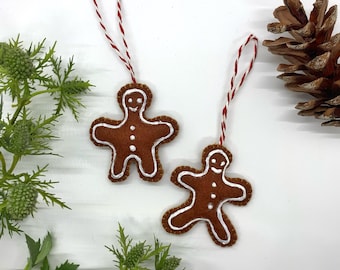 May include: Two felt gingerbread man ornaments with white icing details and smiling faces. Each ornament has a red and white twine hanger.