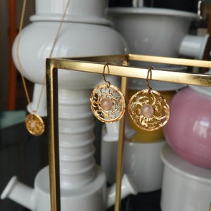 Puede incluir: Pendientes de filigrana dorados con una piedra rosa central, exhibidos en un soporte dorado. También se ve un collar con colgante a juego. Los pendientes y el collar presentan un diseño calado y ornamentado.