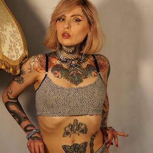 Könnte beinhalten: Ein zweiteiliges Dessous-Set mit Leopardenmuster. Der BH hat dünne schwarze Träger und das Höschen ist ein passender Slip. Das Model hat mehrere Tätowierungen an Armen, Brust, Bauch und Beinen. Eine silberne Halskette wird getragen.