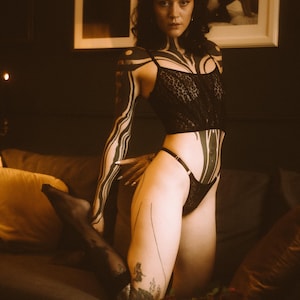 May include: A woman with black and white tattoos poses on a couch wearing a black lace top and matching underwear. She has dark hair and is wearing black stockings.