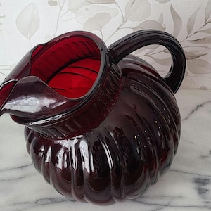 May include: A ruby red glass pitcher with a ribbed, rounded body and a curved spout. The handle is a solid, dark red. The pitcher is likely used for serving beverages and has a vintage aesthetic.