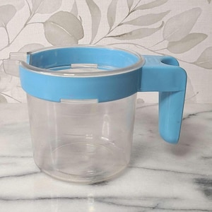 May include: A clear plastic measuring pitcher featuring a bright blue rim and a matching blue handle. The pitcher has a wide circular opening with a small pouring spout on one side and a sturdy, vertical handle attached to the blue rim.
