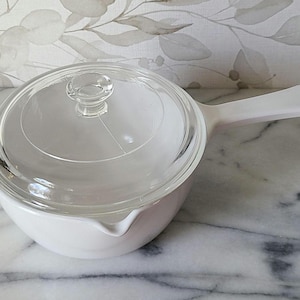 May include: A white, round casserole dish with a clear glass lid and a handle. The dish has a pouring spout and is sitting on a marble surface. The lid has a small knob for easy handling. The overall design is simple and functional.
