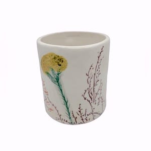 May include: A white ceramic tumbler with a floral design. The tumbler features a yellow flower with a green stem and brown and pink sprigs. The interior of the tumbler is white.