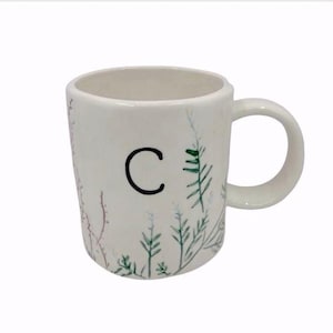 Pu&ograve; includere: Tazza in ceramica bianca con la lettera nera "C" e illustrazioni botaniche ad acquerello in verde e rosso. La tazza ha un manico arrotondato e un design semplice, per bevande calde.