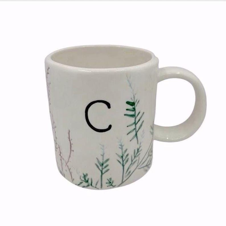 Pu&ograve; includere: Tazza in ceramica bianca con la lettera nera "C" e illustrazioni botaniche ad acquerello in verde e rosso. La tazza ha un manico arrotondato e un design semplice, per bevande calde.