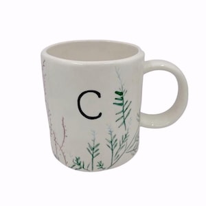 May include: White ceramic mug featuring the letter "C" in black, with painted botanical details in green, blue, and red. The mug has a rounded handle and a simple design, ideal for hot drinks.