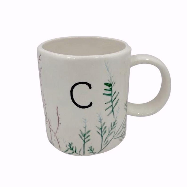 May include: White ceramic mug featuring the letter "C" in black, with painted botanical details in green, blue, and red. The mug has a rounded handle and a simple design, ideal for hot drinks.