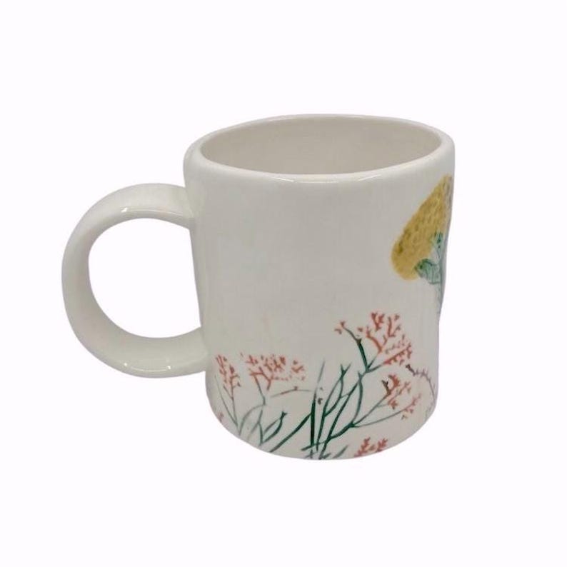 May include: White ceramic mug with a rounded handle. The mug features a floral design with red, green, and yellow accents. The design includes a yellow mushroom and delicate red flowers with green stems.
