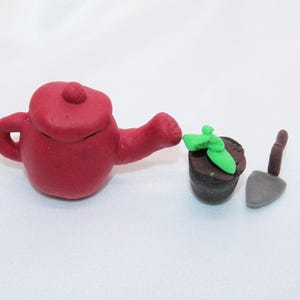 May include: A miniature gardening set made of red, brown, and green clay. The set includes a red watering can, a small pot with a green plant, and a gray trowel. The items are arranged on a white surface.