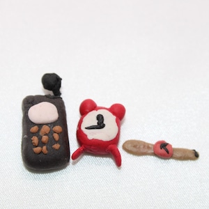 May include: A set of small clay models: a dark brown mobile phone with a white screen, a red alarm clock, and a tan wristwatch with a red face. The items are arranged on a white surface.