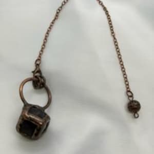 May include: A copper-colored purse charm with a small, square, textured pendant and a connecting chain. The chain ends with a small, round bead. The charm is displayed against a white background.