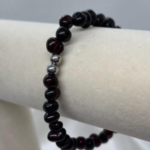 May include: A beaded bracelet featuring dark red, rounded beads. The bracelet has a few silver-colored beads interspersed among the red beads. The bracelet is displayed on a white surface.