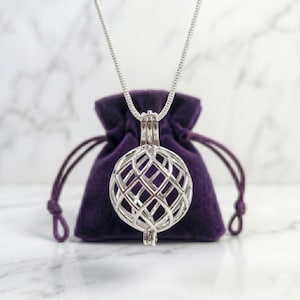 May include: A silver-colored pendant necklace featuring a spherical cage design. The cage is suspended from a delicate chain. The pendant rests against a deep purple velvet pouch, set on a white marble surface.