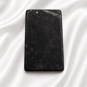 Puede incluir: Una piedra rectangular negra con una superficie ligeramente texturizada. La piedra tiene un aspecto oscuro y moteado y está colocada sobre un fondo blanco y sedoso. Los bordes de la piedra son lisos y bien definidos.