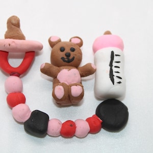 May include: A handmade clay set featuring a brown teddy bear with pink accents, a white baby bottle with a pink top, and a dummy with a red and pink beaded chain. The objects are made of clay.