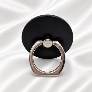 May include: A black phone ring holder with a silver ring. The round base is black, and the ring is silver. The phone accessory is designed to attach to the back of a phone or phone case for a secure grip and stand.