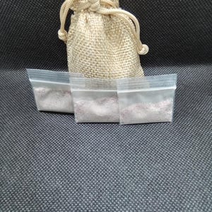May include: A small beige hessian drawstring bag and three small clear plastic bags filled with pink powder. The bags are arranged on a dark grey textured surface. The drawstring bag is tied at the top.