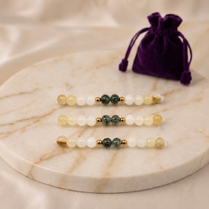 Peut inclure: Trois bracelets de perles présentant un motif symétrique de perles jaune translucide, blanc opaque et vert foncé marbré, agrémentés de petites entretoises métalliques dorées. Une petite pochette à cordon en velours violet est présente.