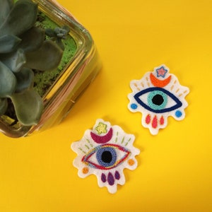 Puede incluir: Dos parches de fieltro bordados con un diseño de ojo malvado colorido. Cada parche presenta un ojo azul con una pupila negra, rodeado de un contorno blanco y acentos coloridos. Un parche tiene una luna creciente, una estrella y acentos naranjas. El otro parche tiene una luna creciente, una estrella y acentos morados.