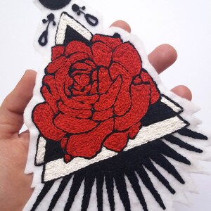 May include: A red embroidered rose with black and white geometric shapes and a crescent moon above it. The rose is surrounded by black and white rays.