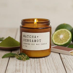 May include: A lit, amber-colored soy wax candle in a glass jar, labeled "Matcha + Bergamot." The candle is 4 oz. and sits on a wooden surface with green matcha powder, limes, and a matchbox.