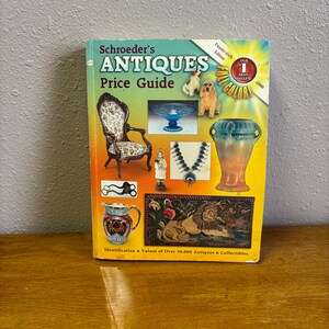 May include: Schroeder's Antiques Price Guide, Twenty-sixth Edition, 2008. A reference book with a yellow and orange cover featuring various antique items including a chair, blue glass bowl, dog figurines, necklace, vase, pitcher, and lion rug.