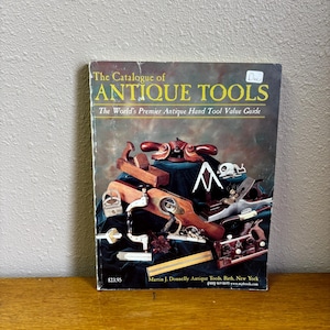 Könnte beinhalten: Ein Buch mit dem Titel "The Catalogue of Antique Tools" mit einem dunklen Einband, der verschiedene antike Handwerkzeuge zeigt. Das Cover des Buches zeigt eine Sammlung von Vintage-Holzbearbeitungswerkzeugen, darunter Hobel und Sägen. Das Buch liegt auf einer Holzoberfläche.