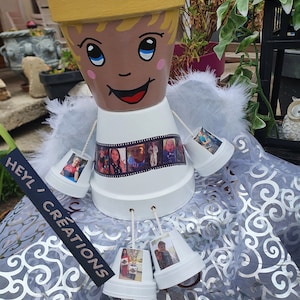 Peut inclure: Un pot de fleurs en céramique blanche en forme d'ange avec une auréole jaune et un visage peint. Le pot a une bande de film avec des photos autour du milieu et est décoré de plumes blanches. Le pot est entouré de plus petits pots en céramique blanche avec des photos qui pendent. Le texte "Heyl Creations" est écrit sur une étiquette noire.