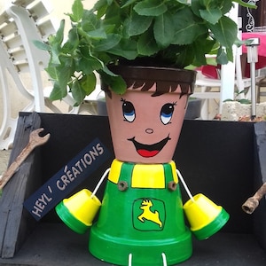 Peut inclure: Un pot de fleurs peint en forme de personne portant une tenue verte et jaune avec un logo John Deere. Le pot est assis sur un banc en bois avec une pancarte qui dit "Heyl' Creations".