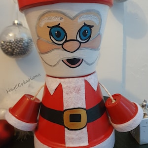 Peut inclure: Un pot en terre cuite peint à la main décoré en Père Noël. Le pot est rouge et blanc avec une ceinture noire et une boucle dorée. Le pot a une barbe blanche et des yeux bleus. Le texte "Hey!l'Creations" est visible sur le pot.