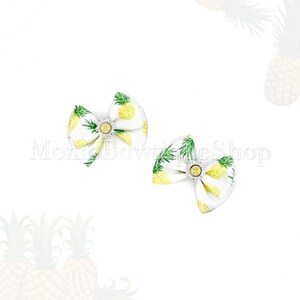 May include: Two white fabric bows with a yellow pineapple pattern and green leaves. Each bow has a silver-colored center with a small, round, gold-colored emblem. The bows are likely for pets or hair accessories.
