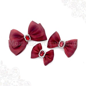 May include: Three burgundy satin ribbon bows with jeweled centers. The bows are different sizes and are arranged in a row on a white background.