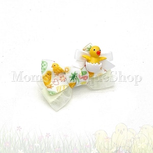 May include: A decorative hair bow featuring a yellow chick emerging from a white eggshell. The bow has layers of white and patterned fabric with yellow chicks and floral designs. The text "Momoboutiqueshop" is visible.