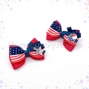 May include: Two patriotic hair bows. Each bow features a red base, a blue ribbon, and a section with the American flag design. One bow has a star charm, and the other has a heart charm with stars and stripes.